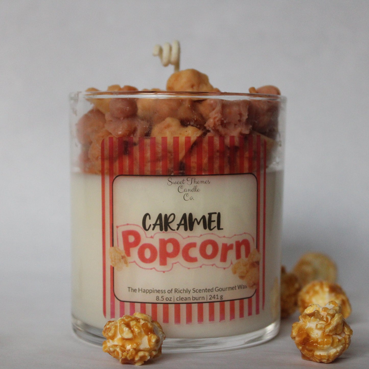 Candle labeled 'Caramel Popcorn' with popcorn kernels and a candle wick on a gray background