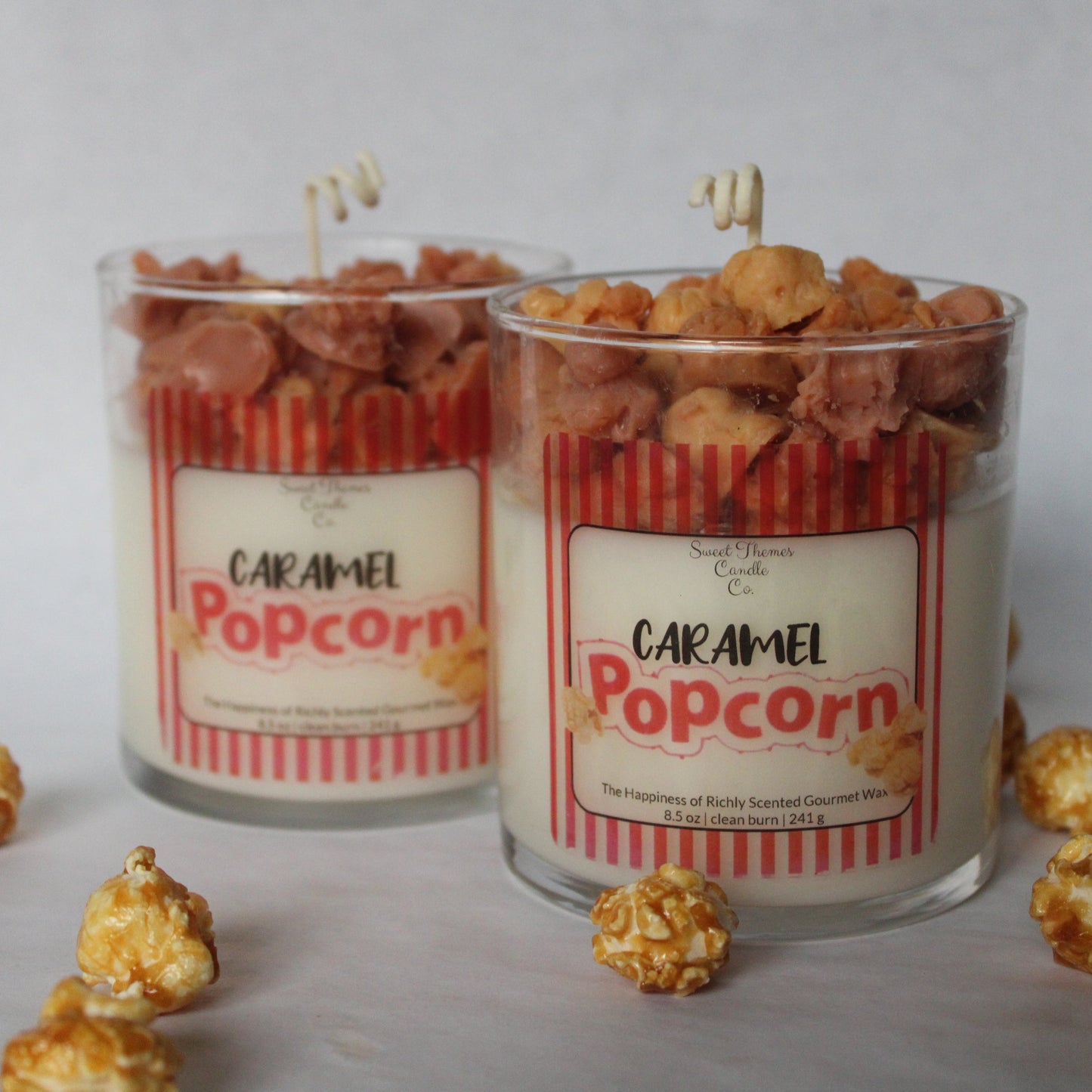 Two 'Caramel Popcorn' candles in glass jars with popcorn scattered around on a light background