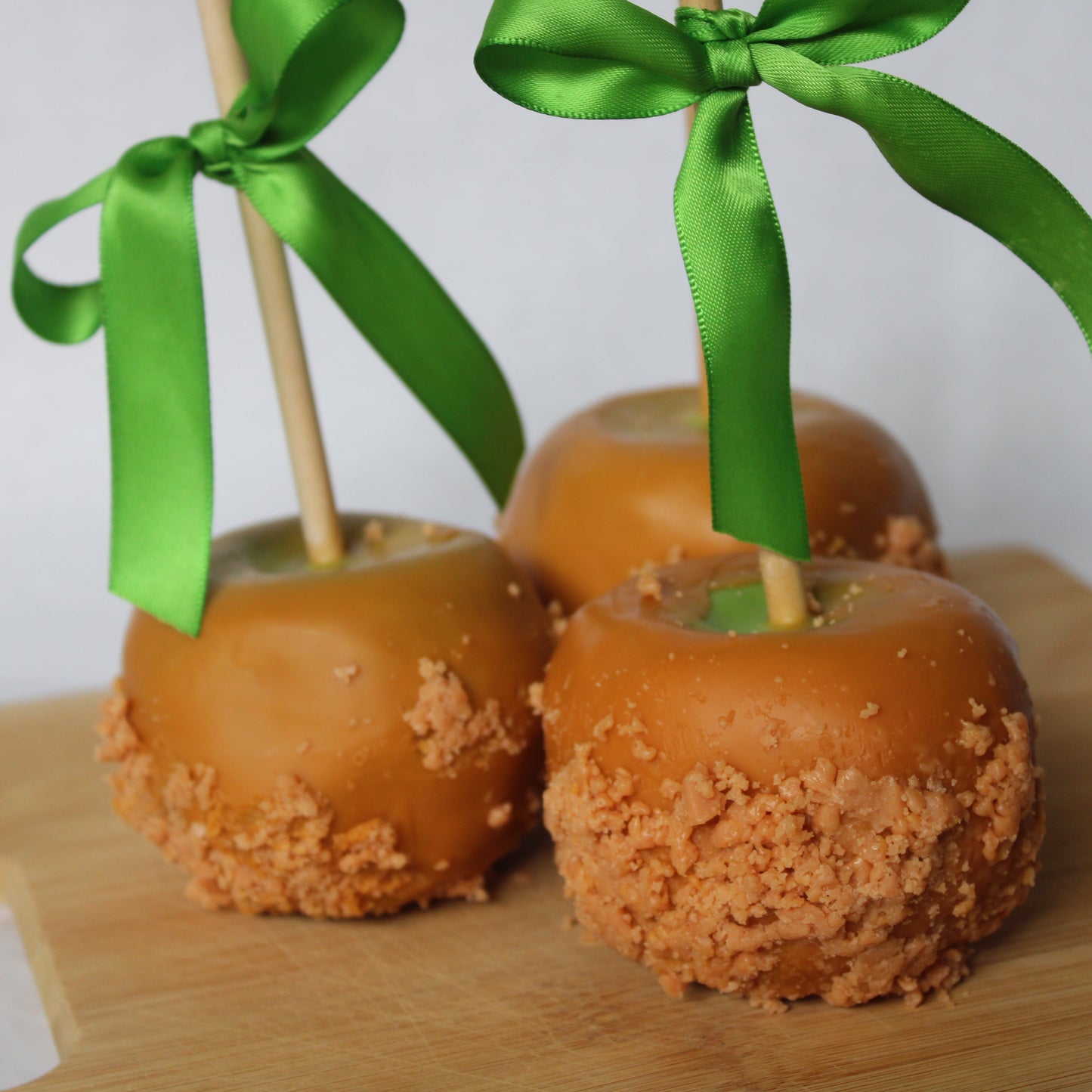 Three caramel apples with green ribbons on a wooden board against a white background