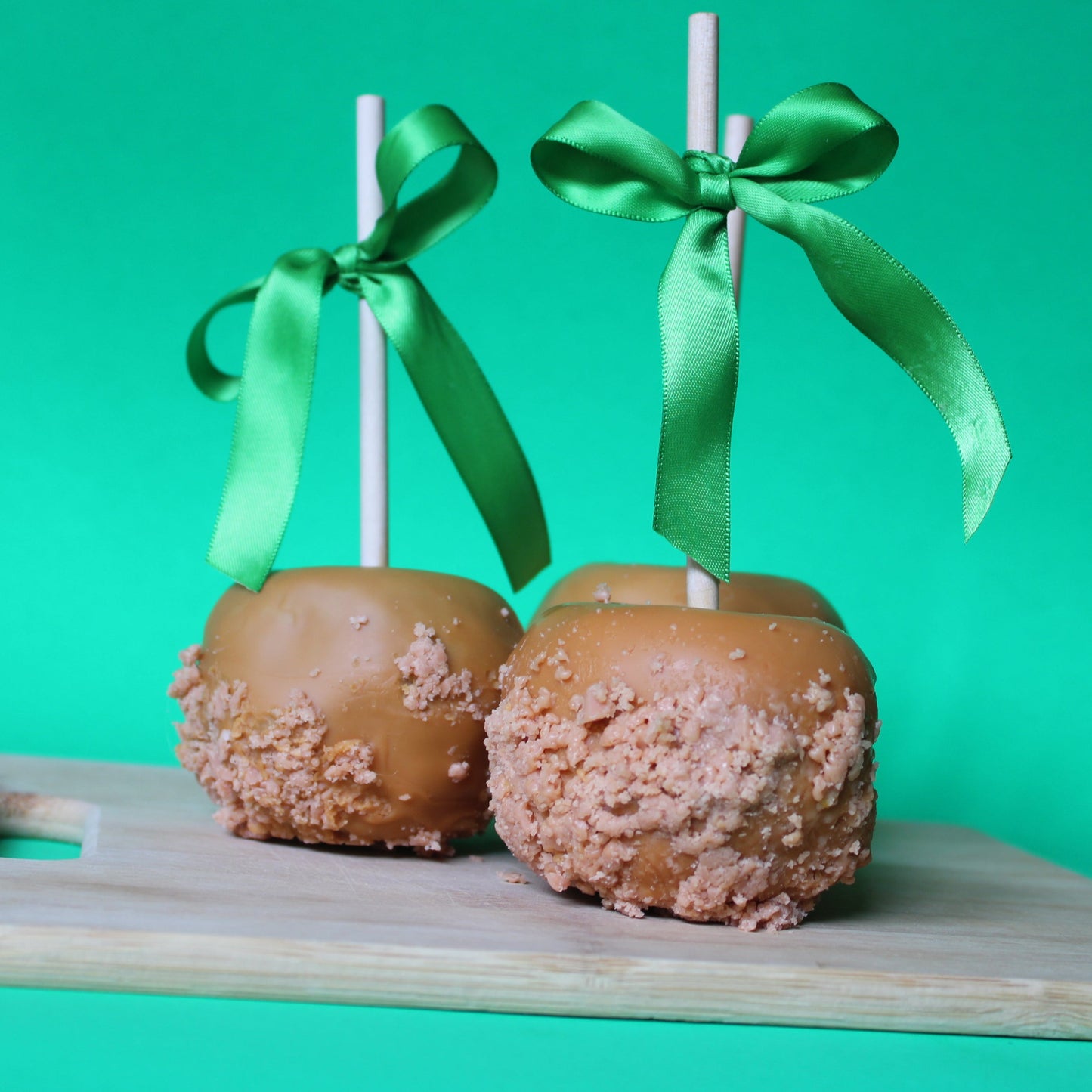Two caramel apples on sticks with green ribbons against a green background
