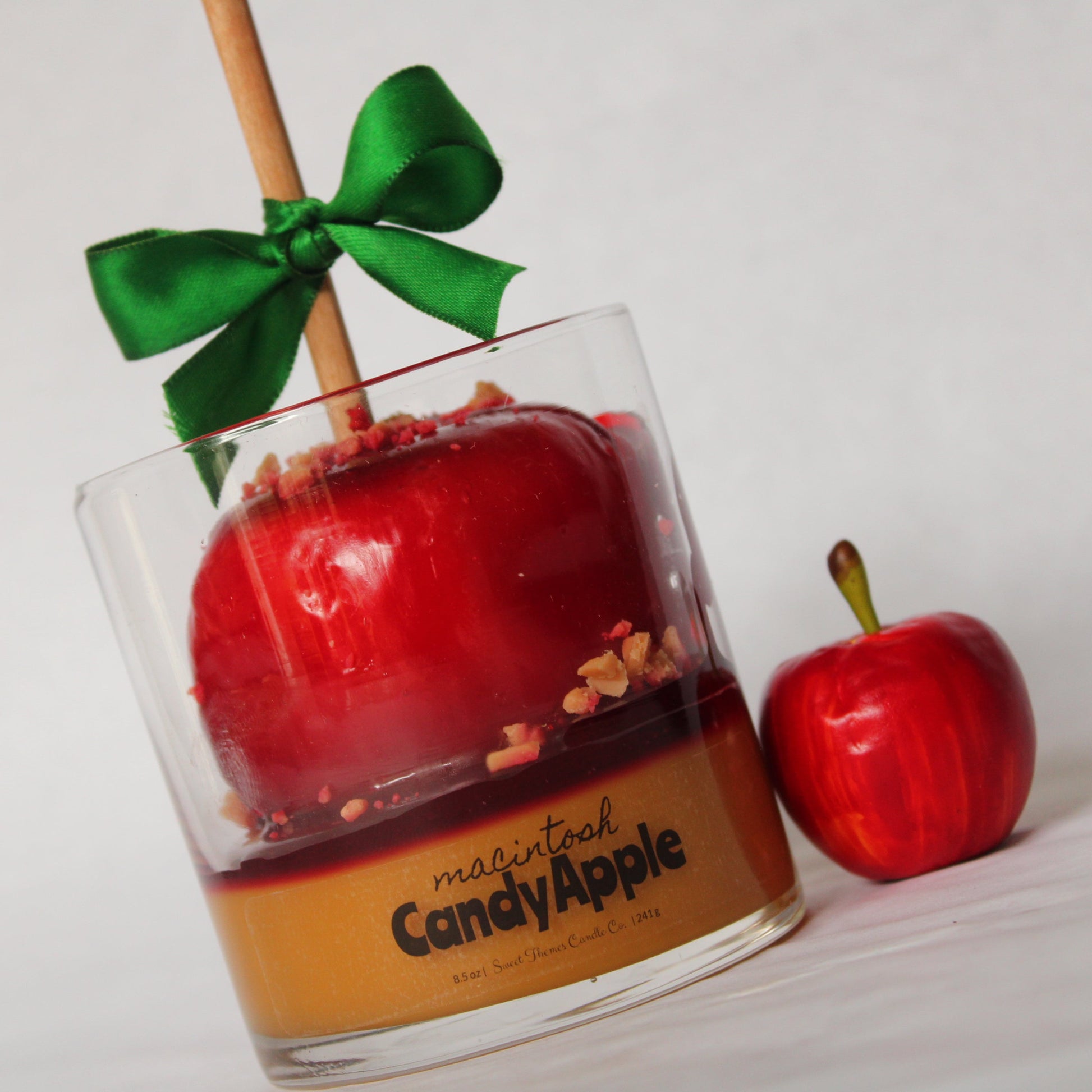 Candy apple in a glass jar with a green ribbon, next to a small red apple on a white background.